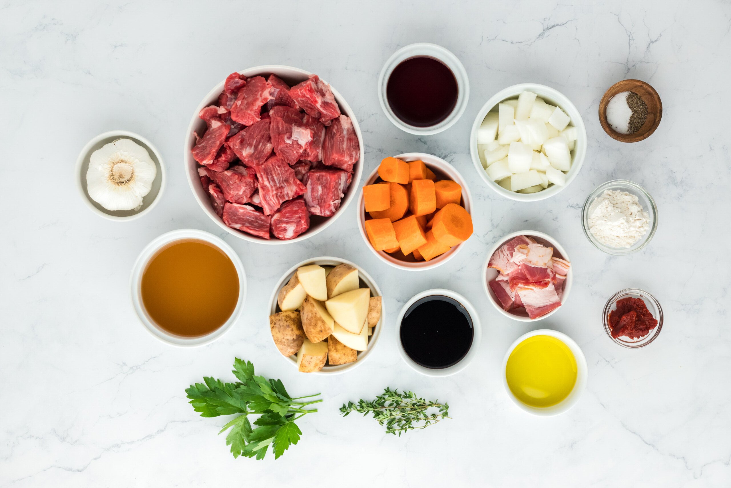 Cubed beef, vegetables, beef stock, herbs, bacon, and spices on a white background in white bowls