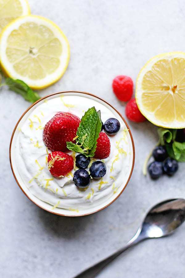Overhead shot of a bowl full of easy lemon mousse topped with fresh berries and mint with lemon slices, raspberries, blueberries and a spoon next to it